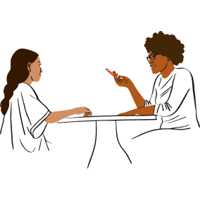 Women sit at table. Woman with brown hair and dark skin speaks and gestures. Woman with brown hair and medium skin listens